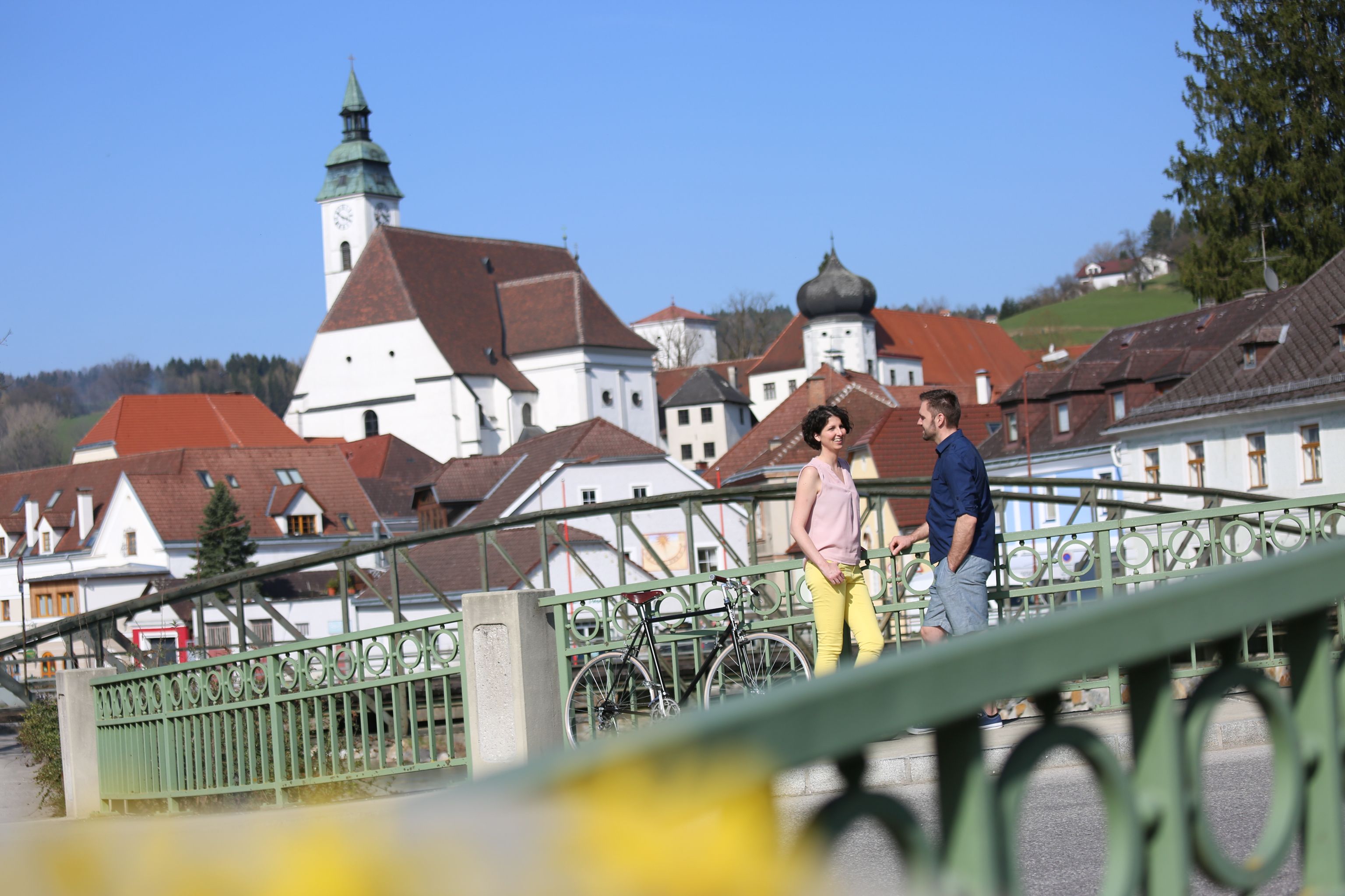 A couple stands on a bridge in Scheibbs with a church in the background.