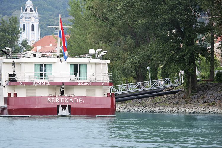 A river cruise ship named Serenade at a landing stage in Dürnstein, with a church tower in the background.