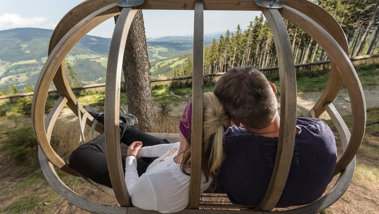 Two people sit in a wooden swing with a view of a hilly landscape.