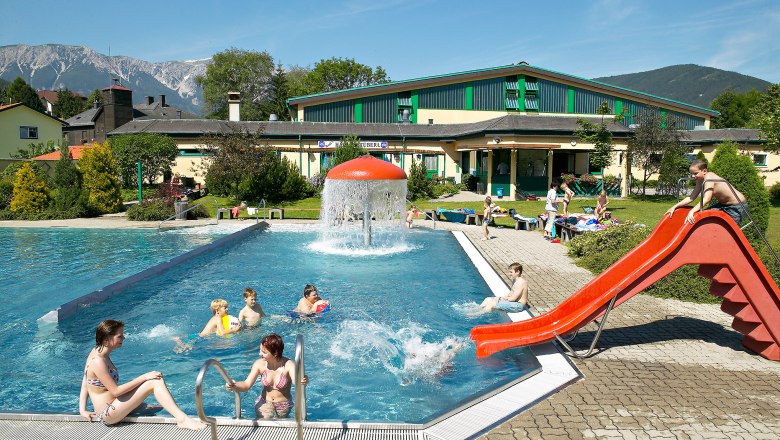 Outdoor pool with slide and mushroom fountain, surrounded by mountains.