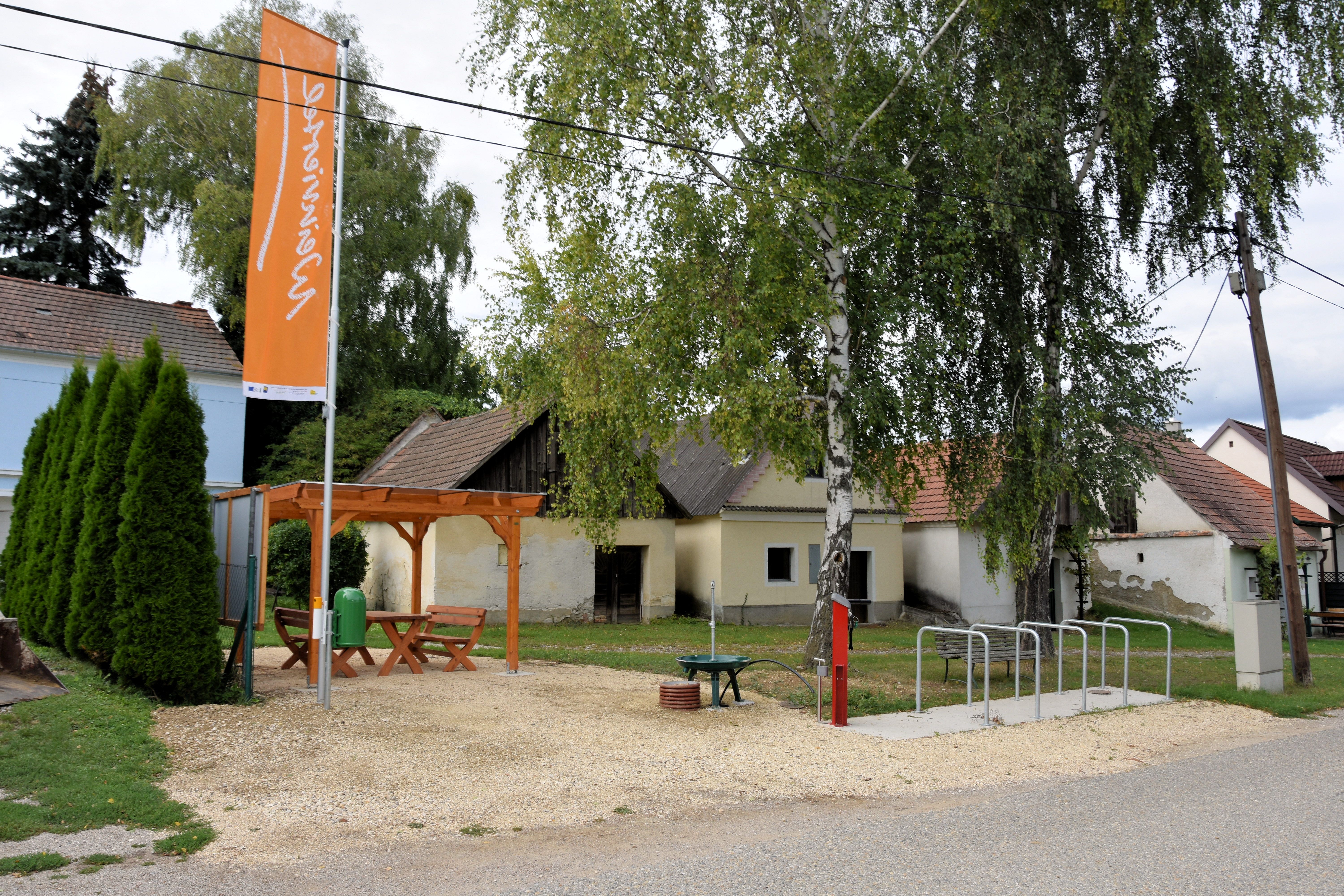 A rest area with a wooden table and benches under a pavilion, surrounded by trees and old buildings. There is also a bike rack and an information sign.