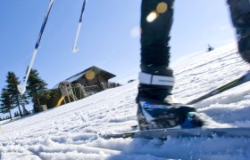 Close-up of a cross-country skier on a snow-covered slope with a wooden hut in the background.