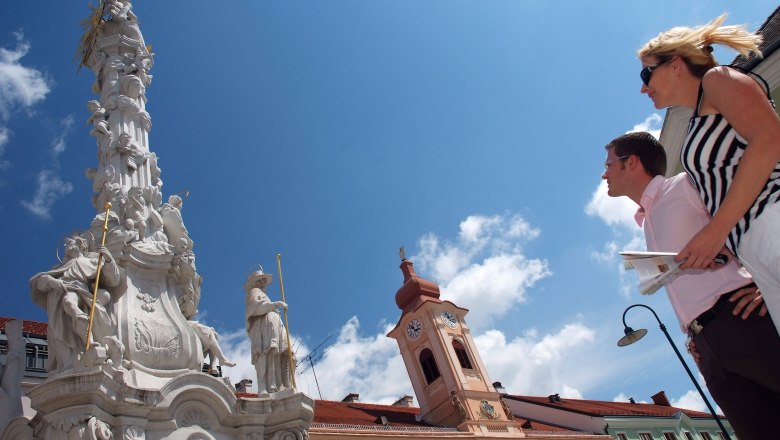 Two people look at a baroque column and a building with a tower under a blue sky.