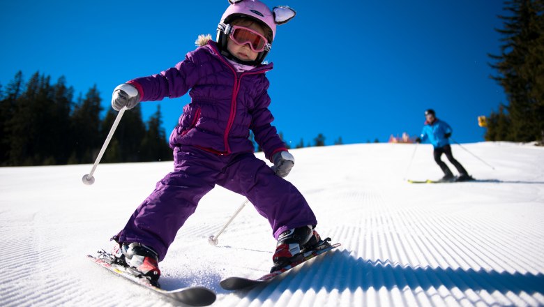 A child in a purple ski suit is skiing down a groomed slope, followed by an adult. The sky is clear and blue.