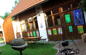A courtyard with wooden tables, posters on a wooden wall and a cauldron over a fireplace.