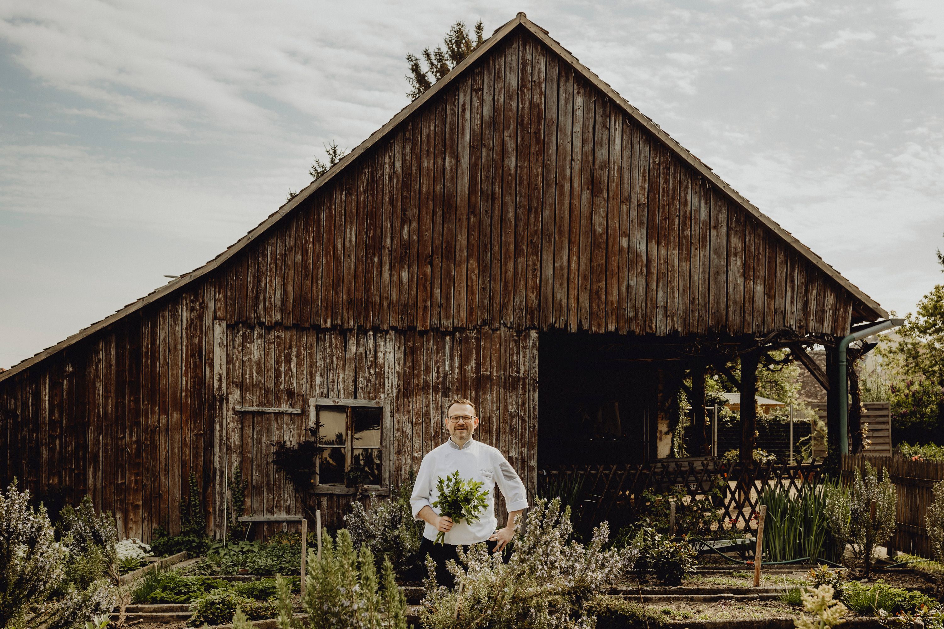 A cook stands in front of an old wooden hut with a bunch of herbs in his hand.
