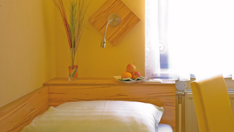 Cozy room with yellow walls, bed, vase with grasses and fruit bowl on a wooden shelf.