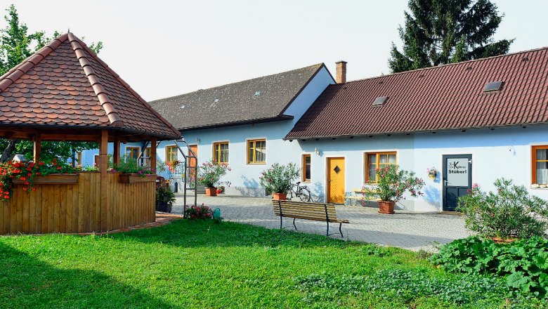 An idyllic courtyard with a pavilion and a blue building with red roofs, surrounded by plants.