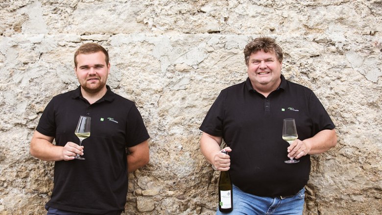 Two men in black polo shirts with wine glasses in front of a stone wall.