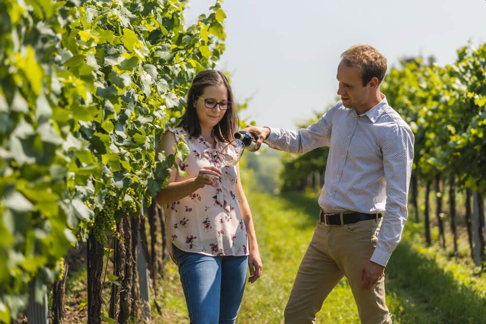 Two people in a vineyard, a woman holding a wine glass, a man pouring wine.