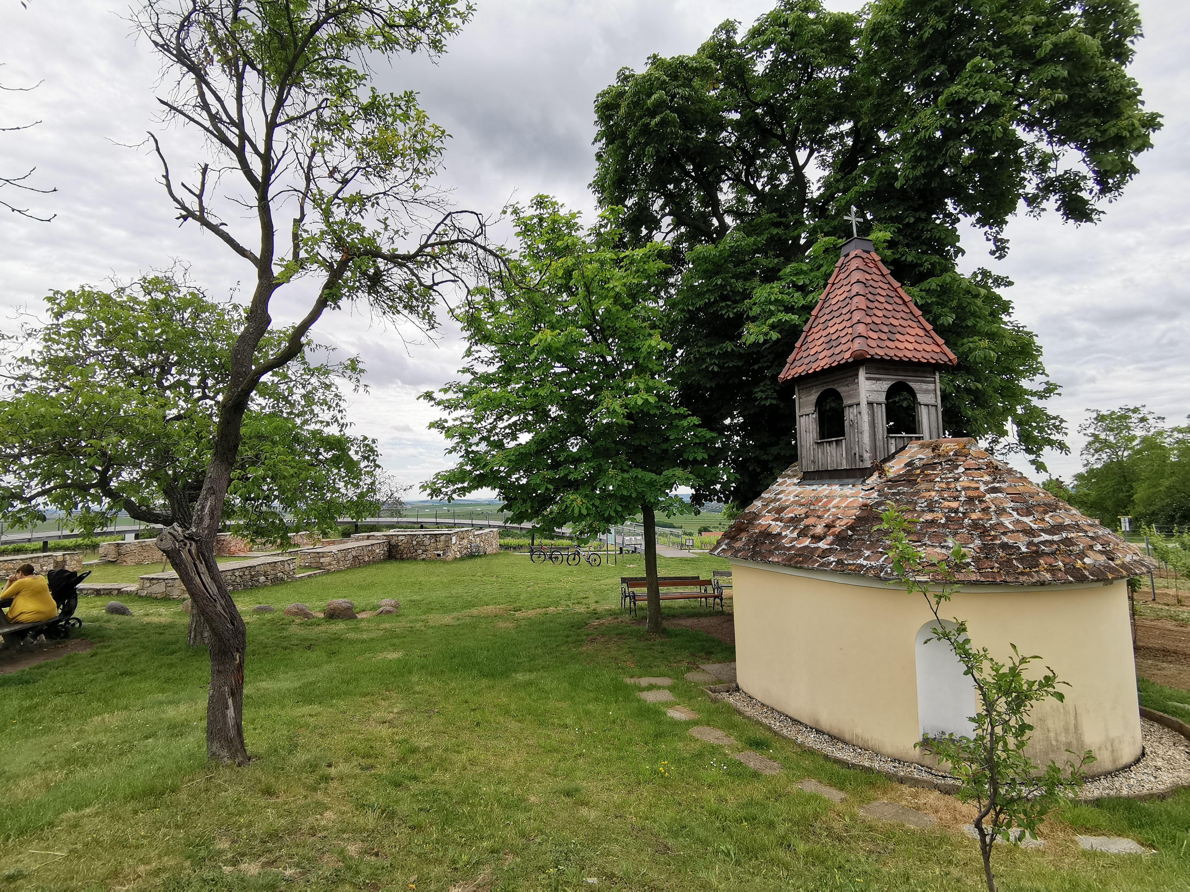 Small chapel with a red tiled roof in a green landscape, surrounded by trees and benches.