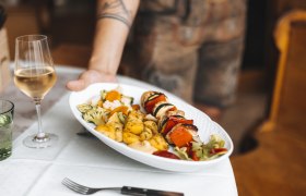 A plate of barbecue skewers, vegetables and potatoes is served, with a glass of white wine next to it.