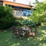 A garden with a wooden table and chairs in front of a vacation home with large windows and surrounded by trees and shrubs.
