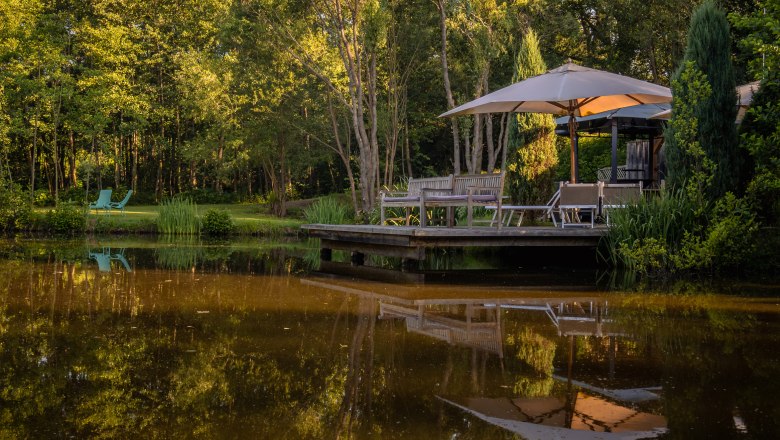 A peaceful pond with trees and a wooden deck with parasol and chairs.