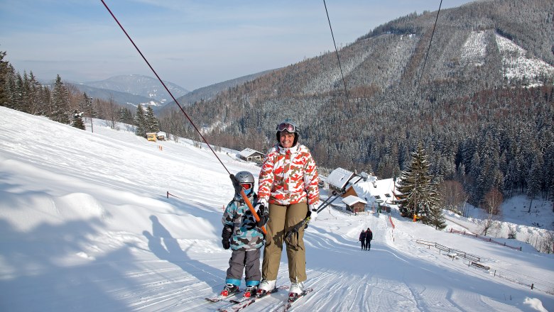 Two skiers on a drag lift in a snowy mountain landscape.
