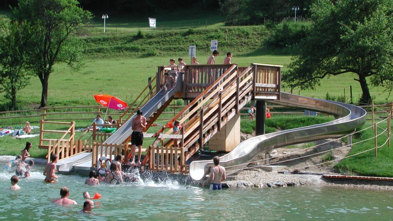 People enjoy a water slide in a natural pool with green surroundings.