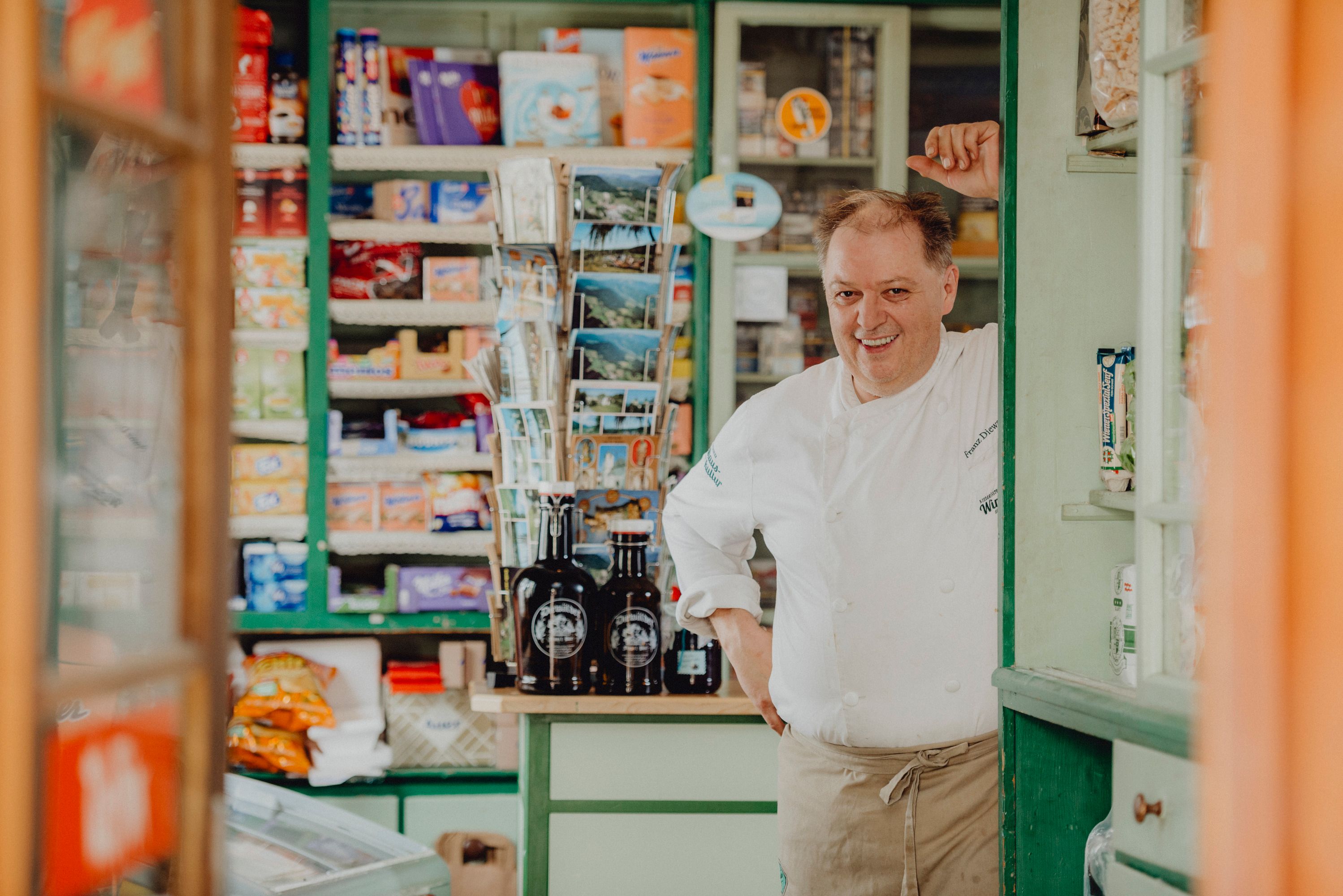 A man in chef's clothes stands smiling in an old store with shelves full of products.