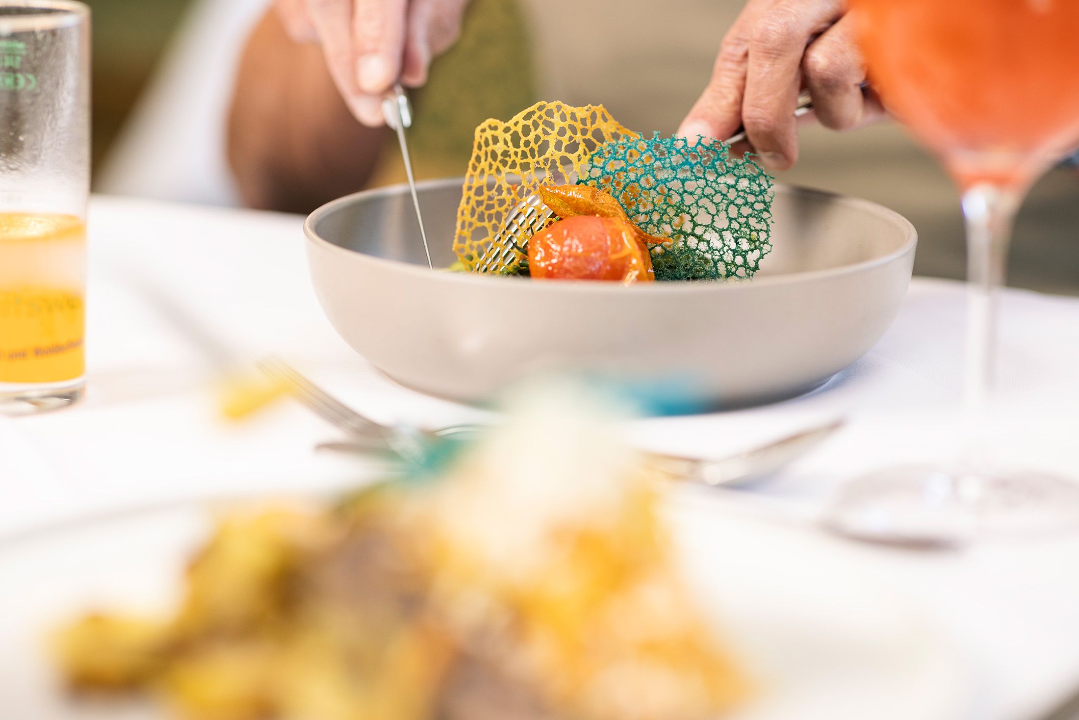 "At Restaurant Kreuzberg, a guest is served a freshly prepared dish at a set table. Drinks and food are on the table. The bright, friendly atmosphere offers a glimpse into the dining experience and the personalized service."