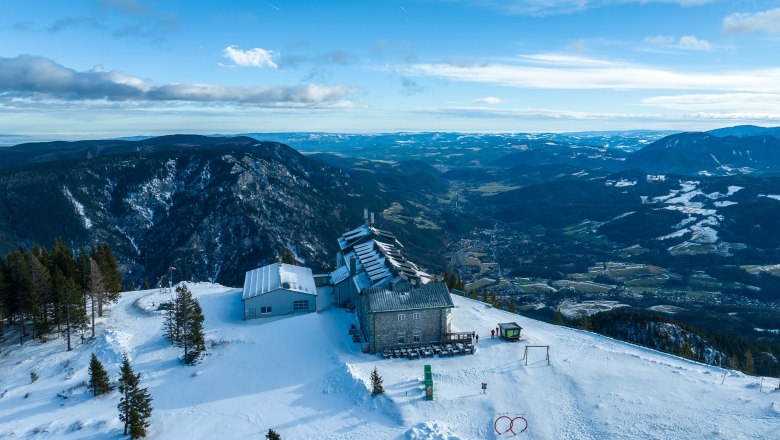Aerial view of the Raxalm mountain inn in winter with snow-covered mountains and valleys in the background.