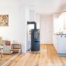 Modern living area with wooden floor, wood-burning stove, dining table and kitchenette.