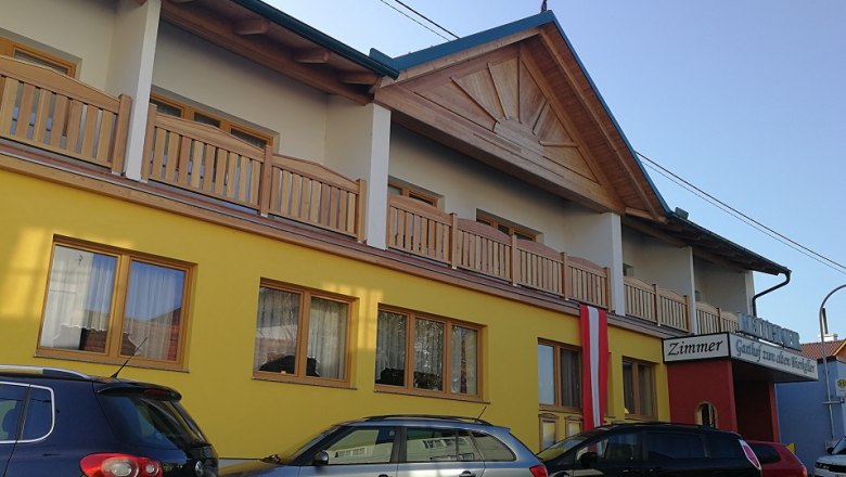 Exterior view of a yellow building with a wooden veranda and cars in front of it.