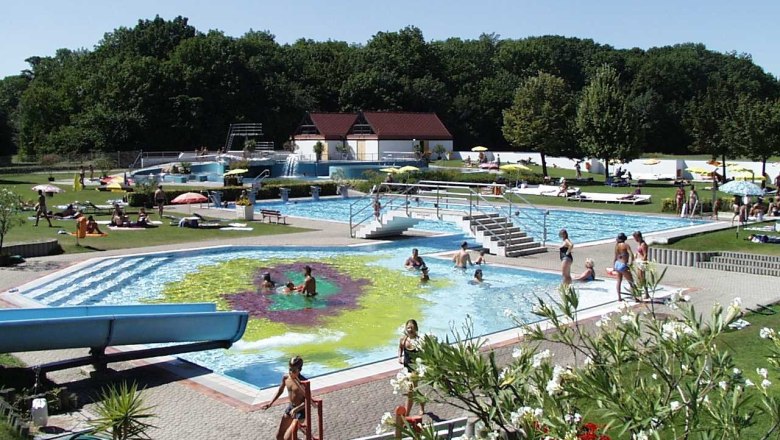 Outdoor pool with several pools, slide and sunbathing lawn, surrounded by trees.