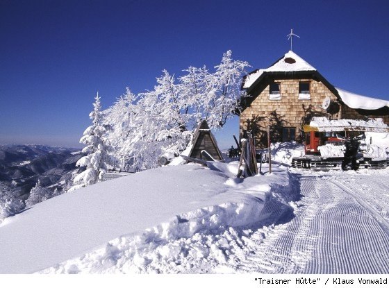 Snow-covered mountain hut with snow-covered trees and a clear blue sky.
