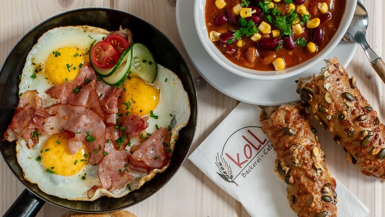 Various hot snacks on a table, including fried egg with bacon, chili con carne, sandwiches and a roll.