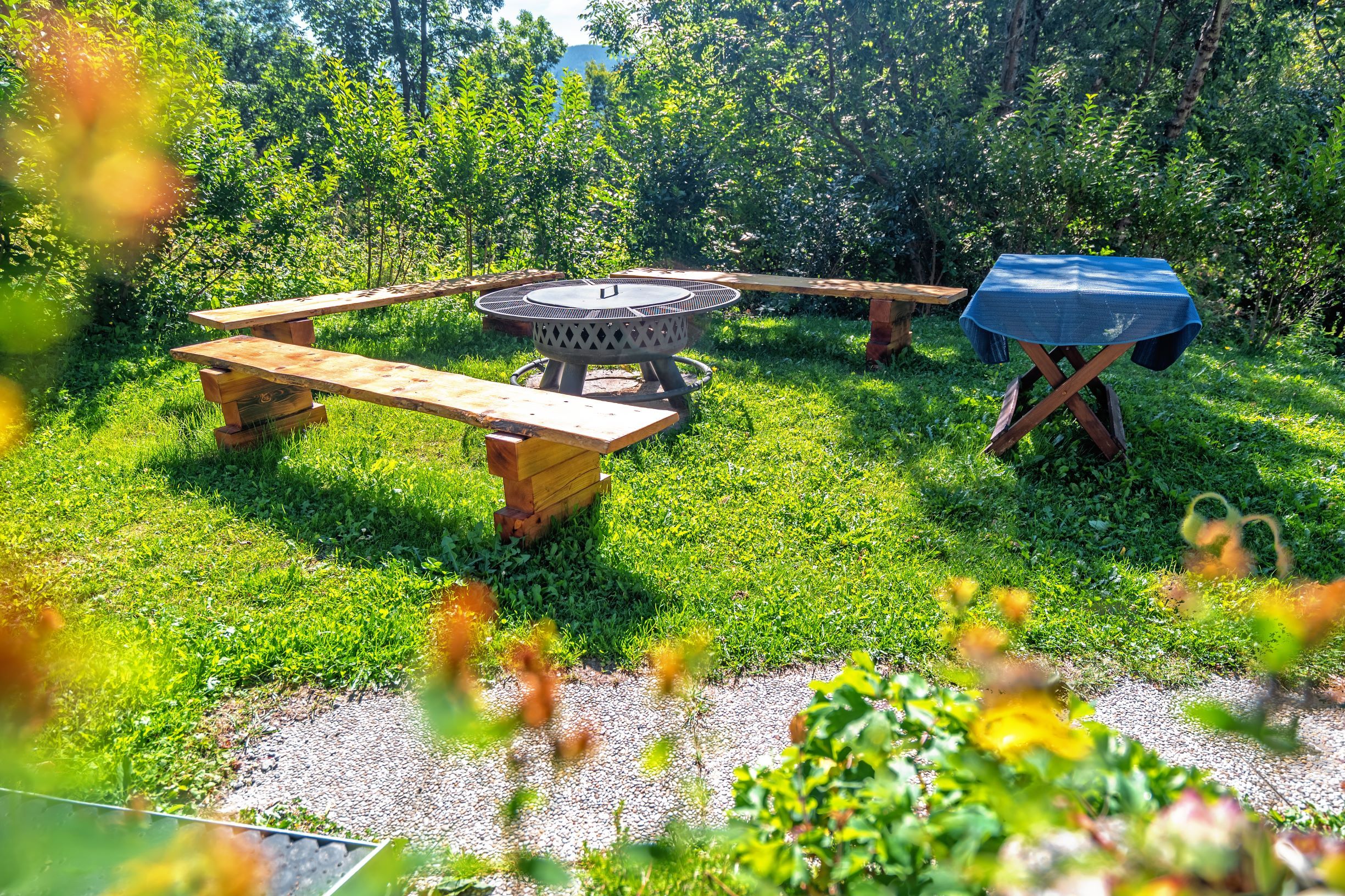 A barbecue area with wooden benches and an outdoor table, surrounded by green vegetation.