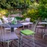 Garden area with tables and chairs on a wooden terrace, surrounded by green plants.