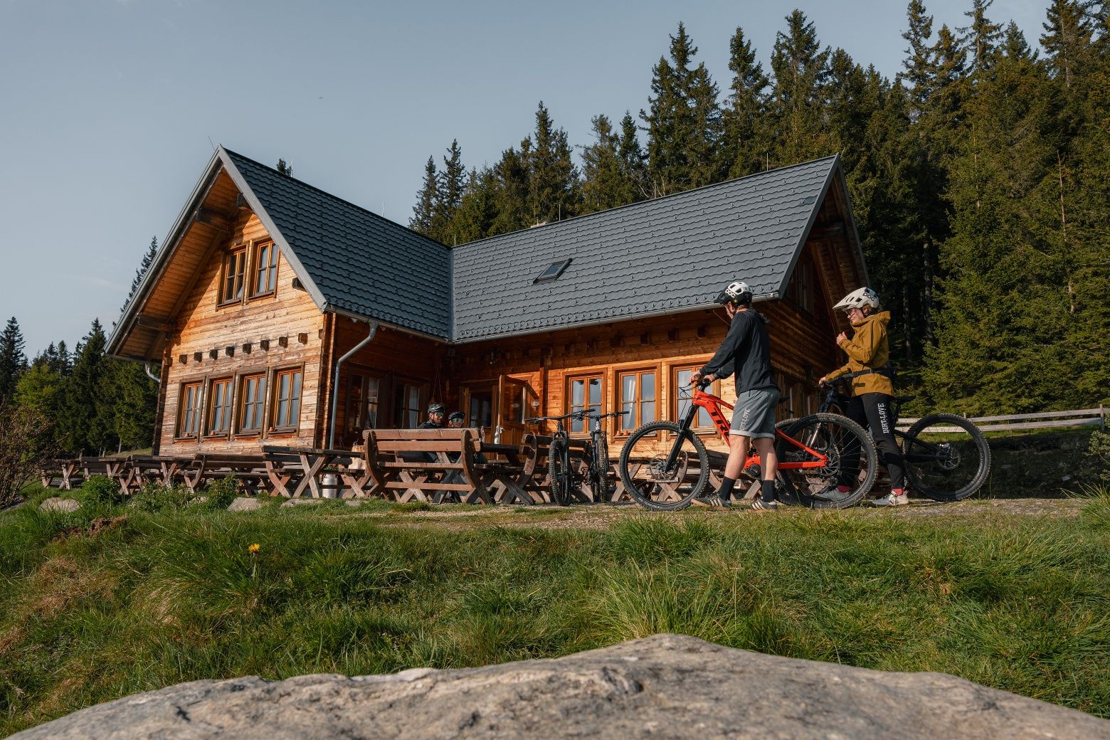 Two mountain bikers are standing in front of a wooden hut in a forest landscape.
