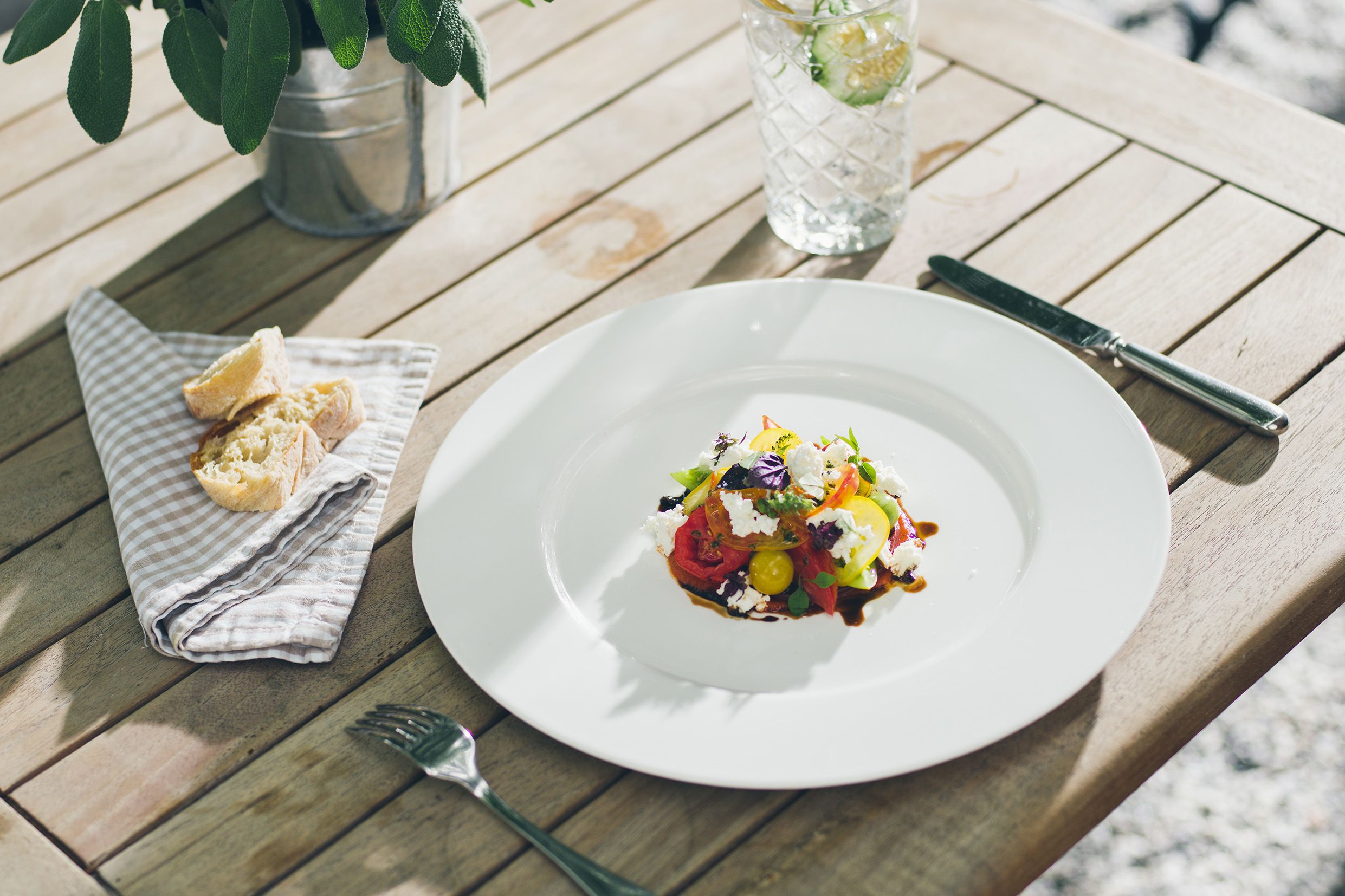 An artfully arranged salad on a white plate, next to it a glass of water with cucumber slices and bread on a napkin.