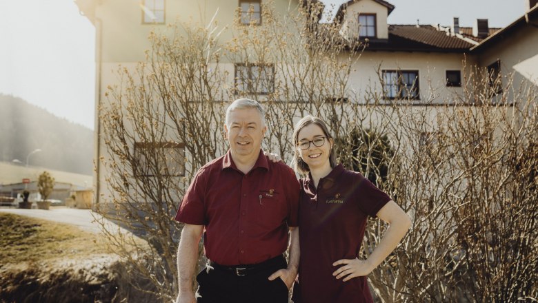 Two people in red shirts stand smiling in front of a building.