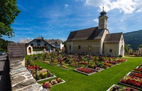 Parish church of St. Peter am Neuwald with a well-tended cemetery and flower beds in bloom, surrounded by a green landscape and blue sky.