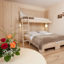 A cozy hotel room with a double bed and a bunk bed, a wooden table with a bouquet of flowers and a closet.