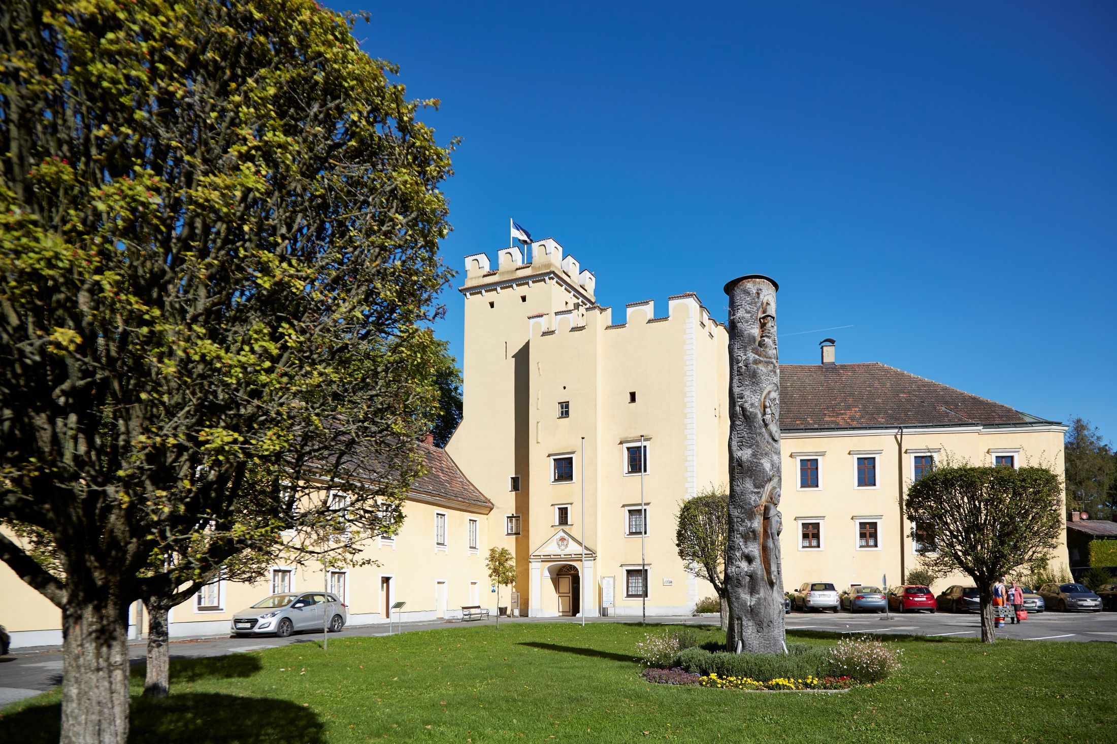 Groß-Siegharts Castle with yellow building and tower, surrounded by trees and a meadow with a sculpture.