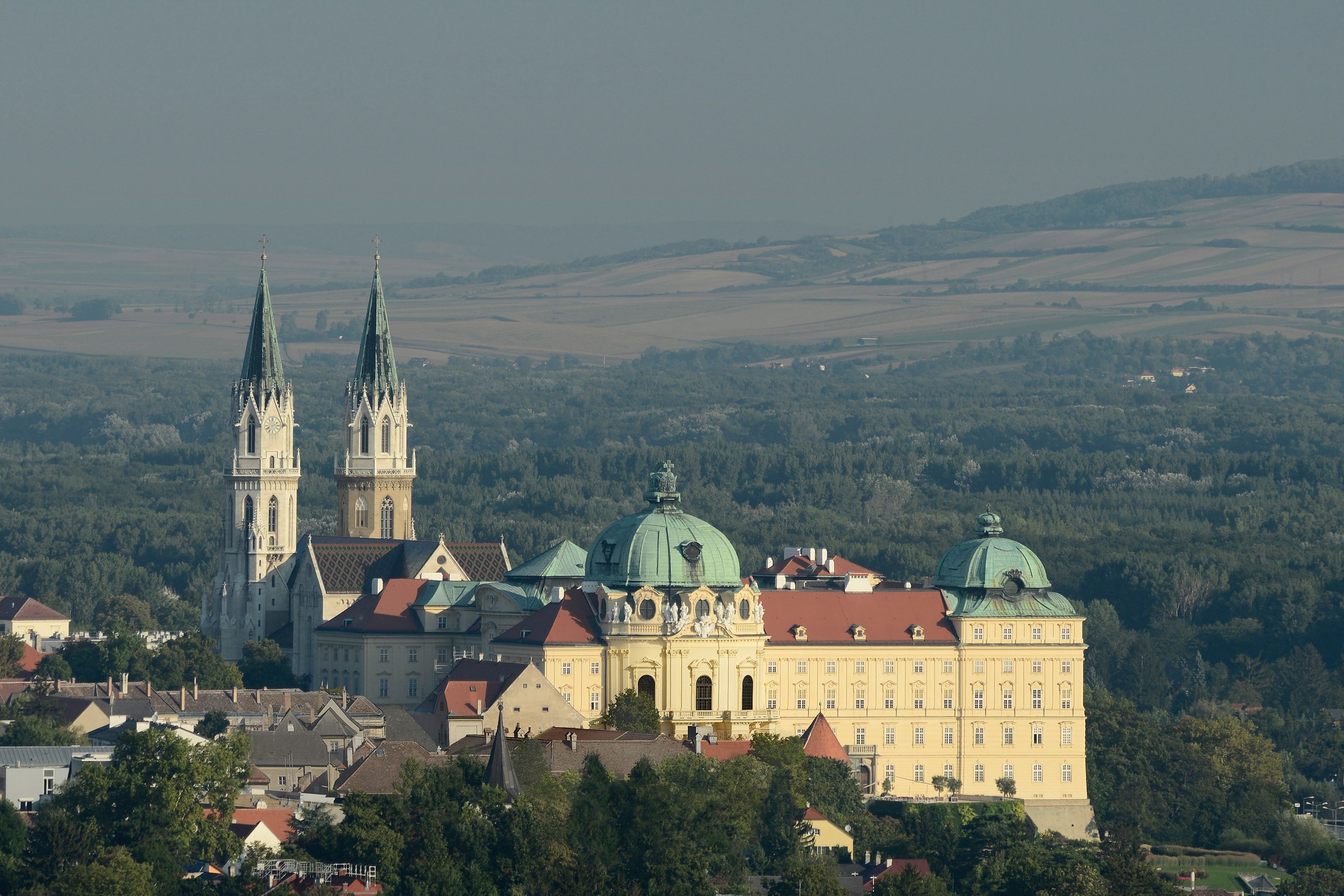 Aerial view of Stift Klosterneuburg Abbey with two towers and a dome, surrounded by forest and fields.