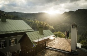 Two people stand on a terrace with a view of wooded mountains at sunset.
