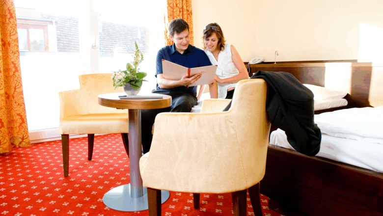A couple is sitting on a bed in a hotel room, looking into a book. The room has red carpets and yellow curtains.