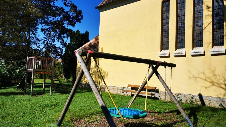 Playground with swing and slide next to a yellow chapel with high windows.