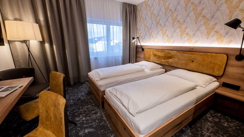 Modern guest room with two single beds, wooden furniture, yellow chairs and a floor lamp. Light-colored curtains and patterned wallpaper on the wall.