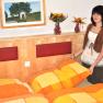 A woman stands in a bedroom with orange and yellow bed linen and vases of flowers on the headboard.