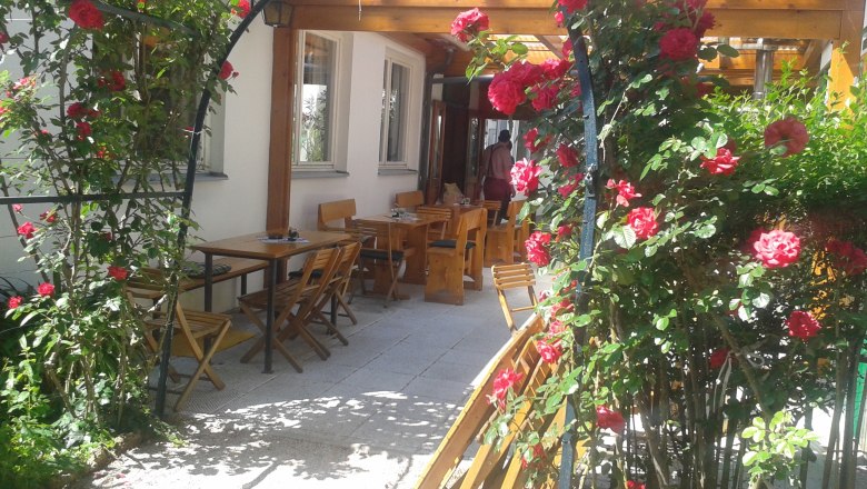 A rose arch with red roses leads to an outdoor area with wooden tables and chairs.
