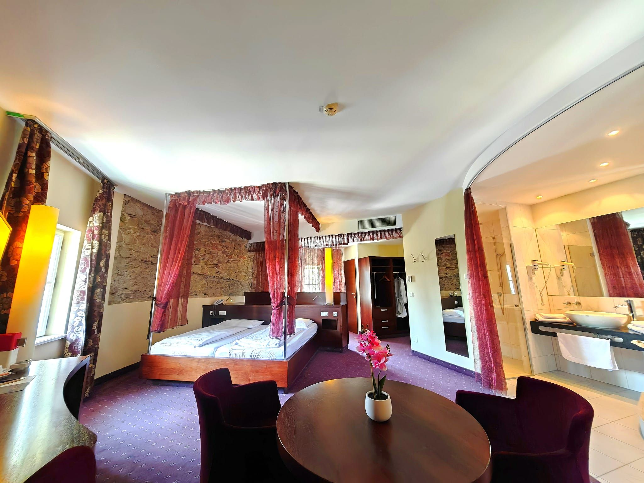 An elegant hotel room with a four-poster bed, a round table with flowers, and an open-concept bathroom area.