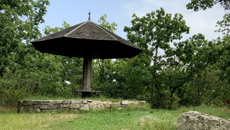 A wooden pavilion with a pointed roof in a green, wooded setting.