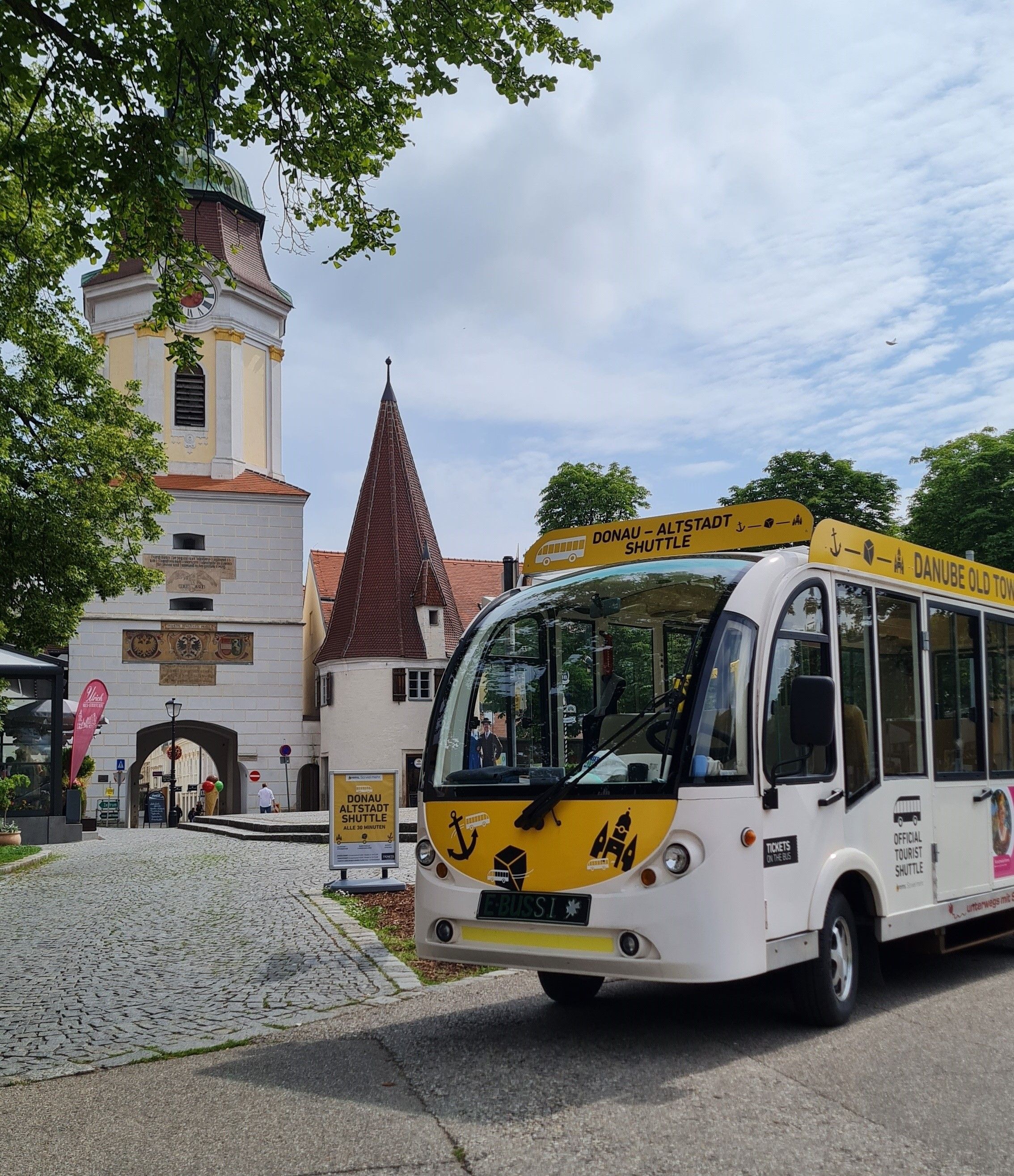 A white old town shuttle bus stands in front of a historic archway and a church tower.