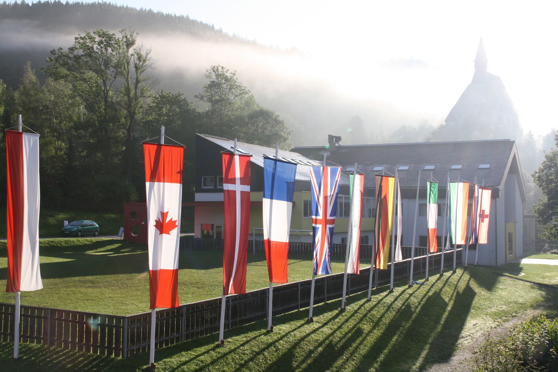 Various national flags in front of a building in a rural setting with fog and trees.