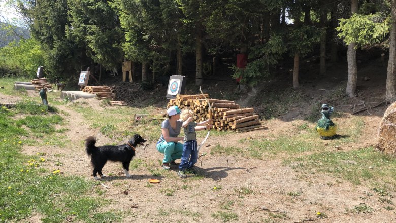 A child and an adult practicing archery outdoors, accompanied by a dog. Targets and trees can be seen in the background.