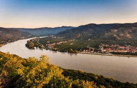 Panoramic view of a river, surrounded by hills and villages, at sunset.