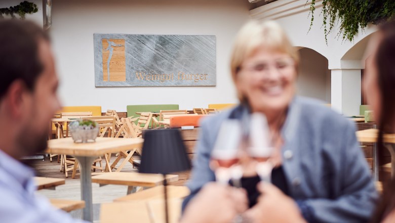Three people clink glasses of wine in a courtyard, with a sign reading 'Weingut Burger' in the background.